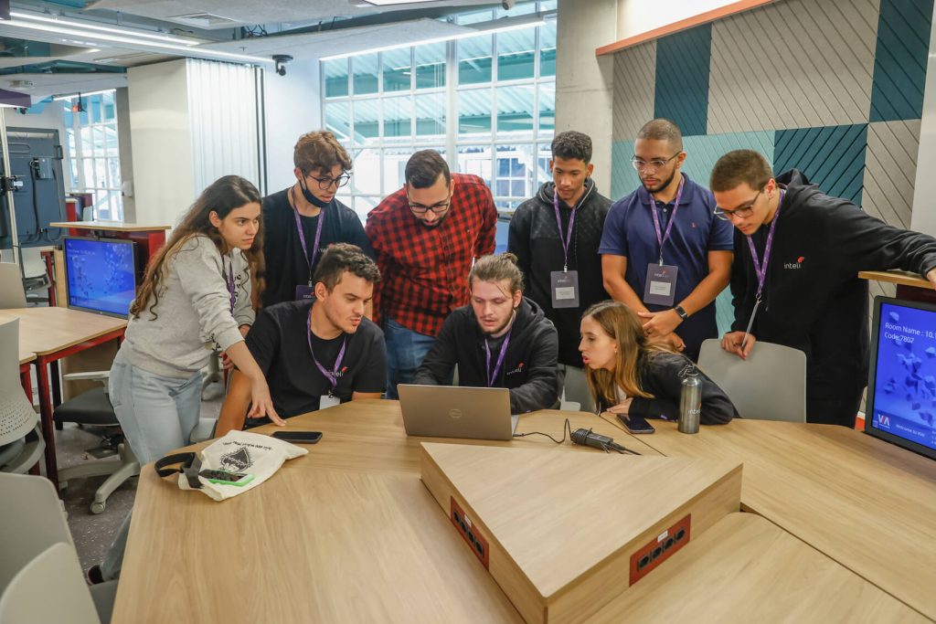 A group of Inteli college students, sitting at desks, are collaborating on a project for a large company. They are engaged in a practical activity, possibly brainstorming or prototyping. The room is modern and well-lit, with equipment and work materials visible on the tables. The atmosphere is one of concentration and teamwork, with the students actively interacting with each other.